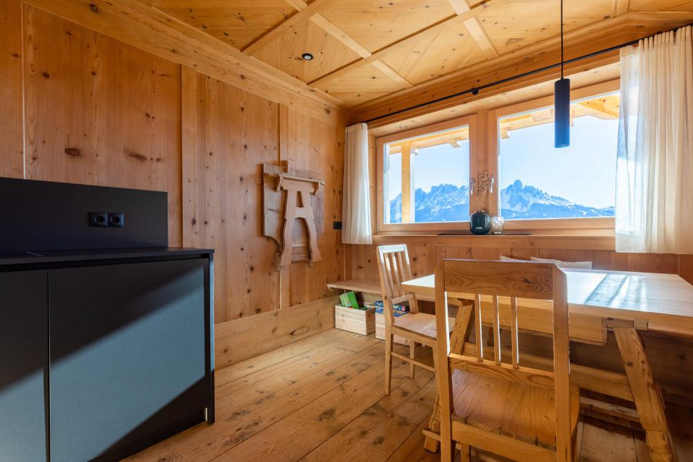 Wood-panelled living room with a dining table, wooden chairs and a bench by the window, view of the Dolomites in the background, dark kitchenette on the left Wood-panelled living room with a dining table, wooden chairs and a bench by the window, view of the Dolomites in the background, dark kitchenette on the left
